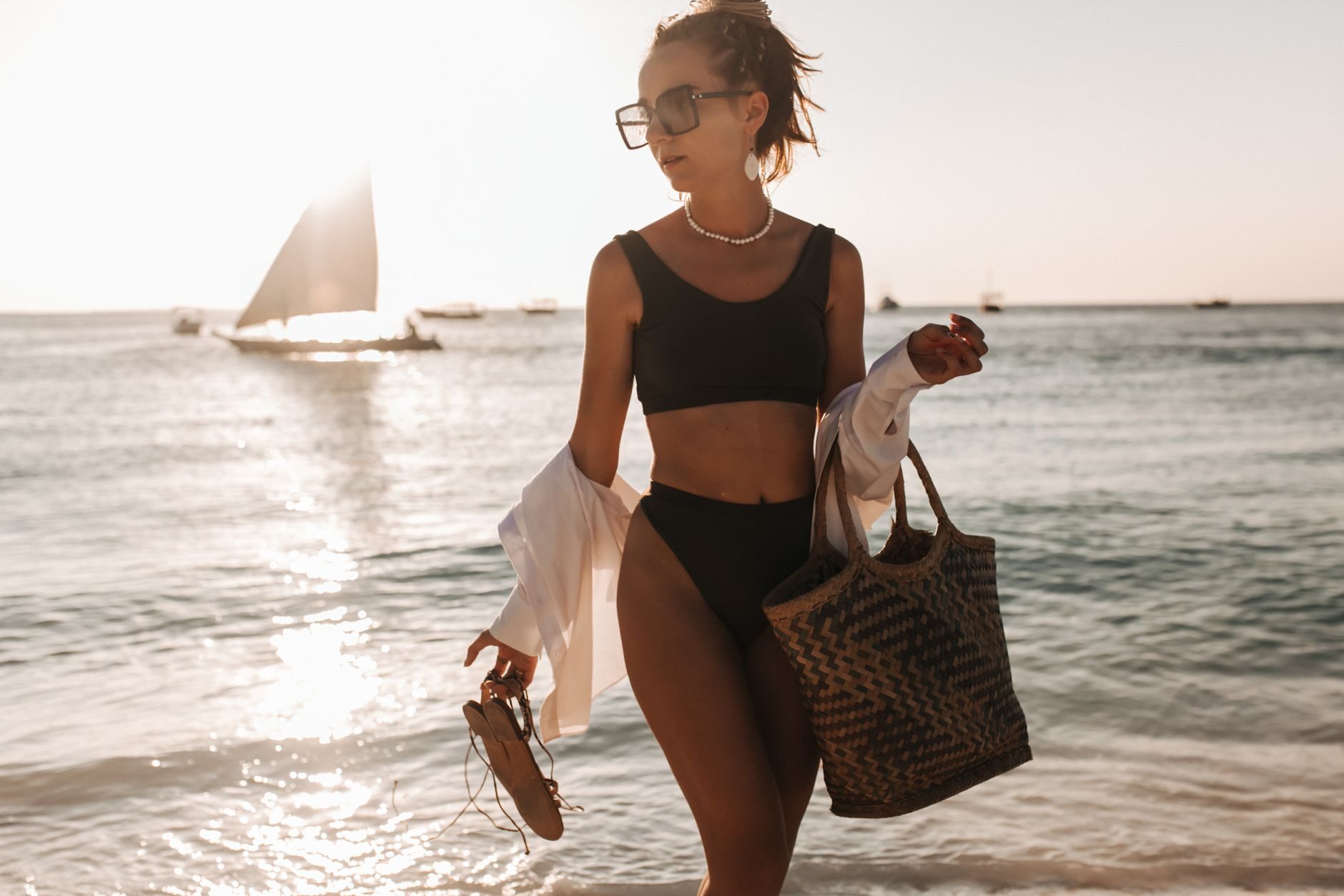 A confident woman walking along a turquoise Gulf shoreline at golden hour, sailboats behind her